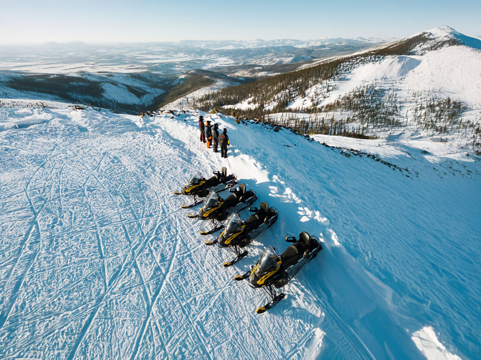 Aerial view of four people and four Ski-Doo snowmobiles parked on a snowy mountain ridge under a clear blue sky.