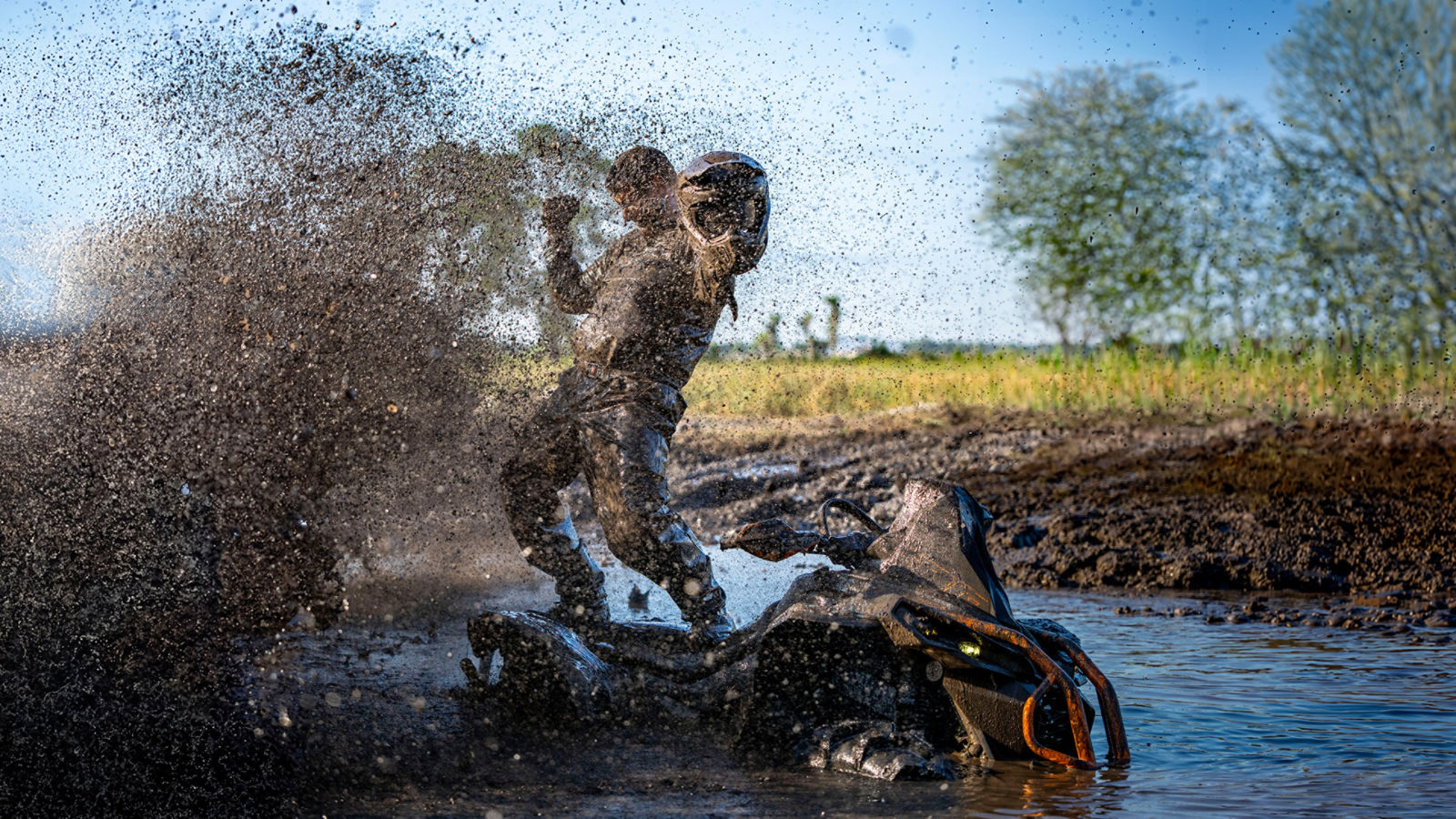 A muddy rider standing triumphantly on a Can-Am ATV in a large splash of water and mud.