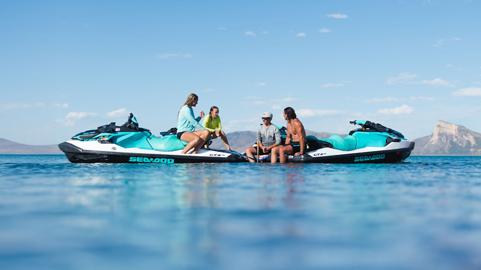 Four people relaxing on four Sea-Doo personal watercraft tied together on calm blue water with mountains in the background.
