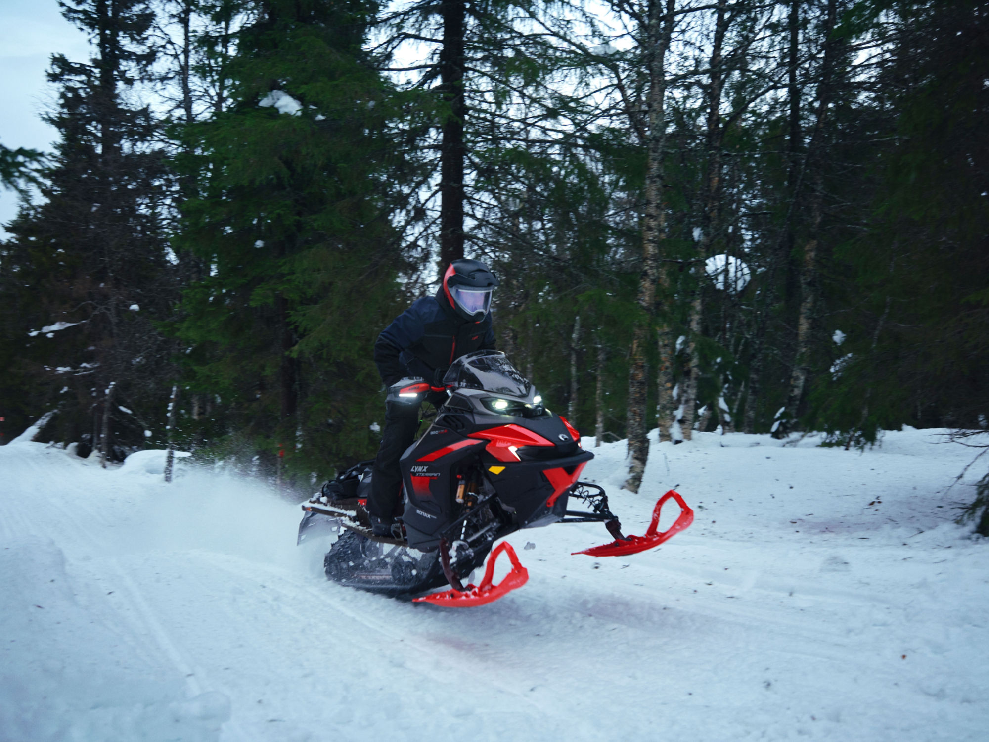 2025 Lynx Xterrain snowmobile riding fast on a snow-covered trail through a forest.