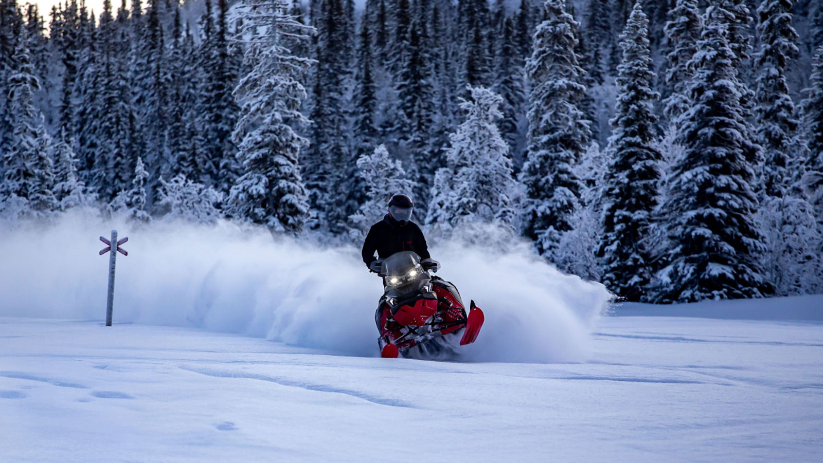 A snowmobiler riding the 2026 Lynx Brutal snowmobile through the deep snow of a snow-covered forest.