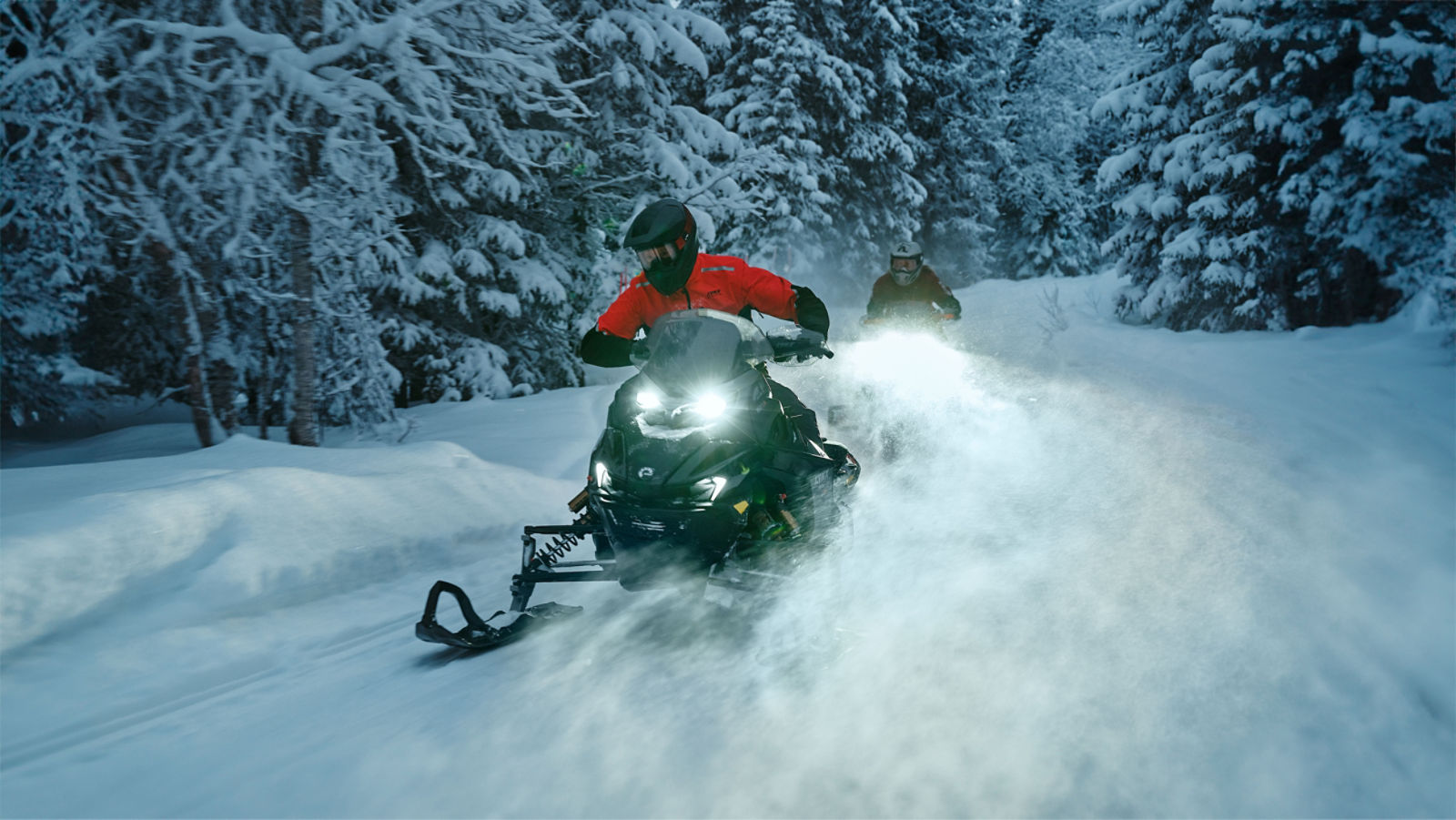 Deux motoneigistes sur leur motoneige Lynx Rave 2026 traversant à toute vitesse une piste de forêt enneigée, en soulevant de la poudreuse fraîche.