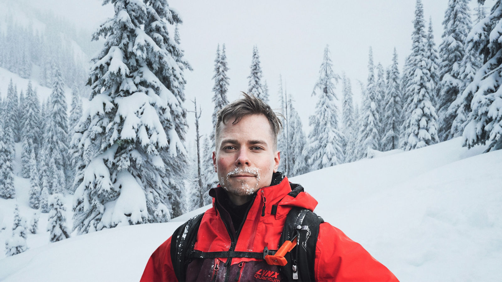 En man står i snöskoterutrustning i ett snöigt landskap, med rimfrost i skägget