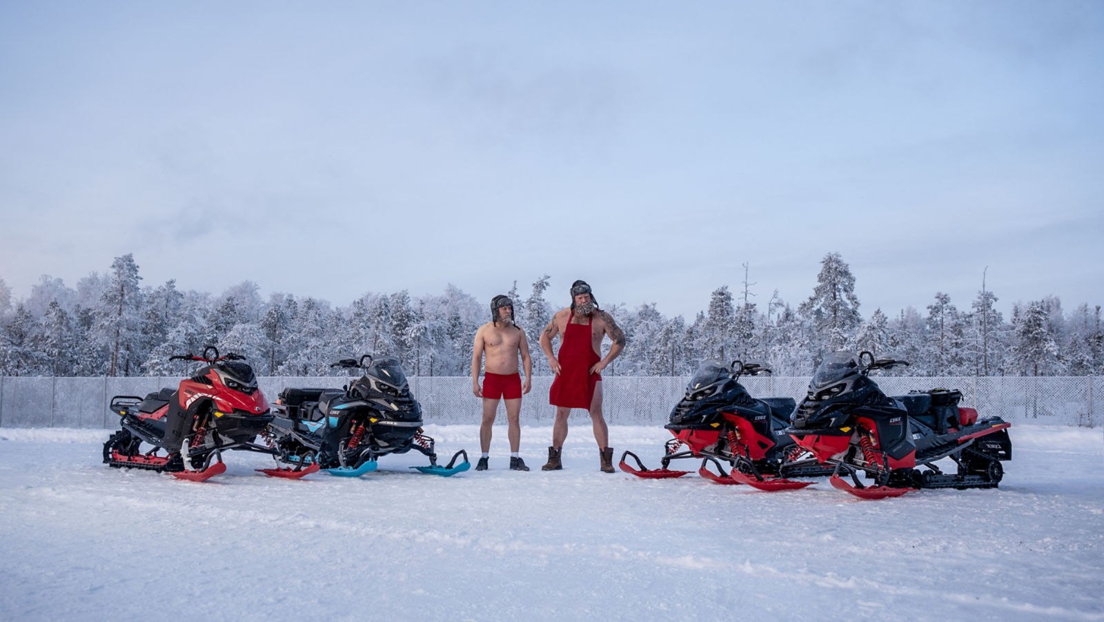 Två män med pälsmössor står mitt bland fyra Lynx-snöskotrar på en gård.