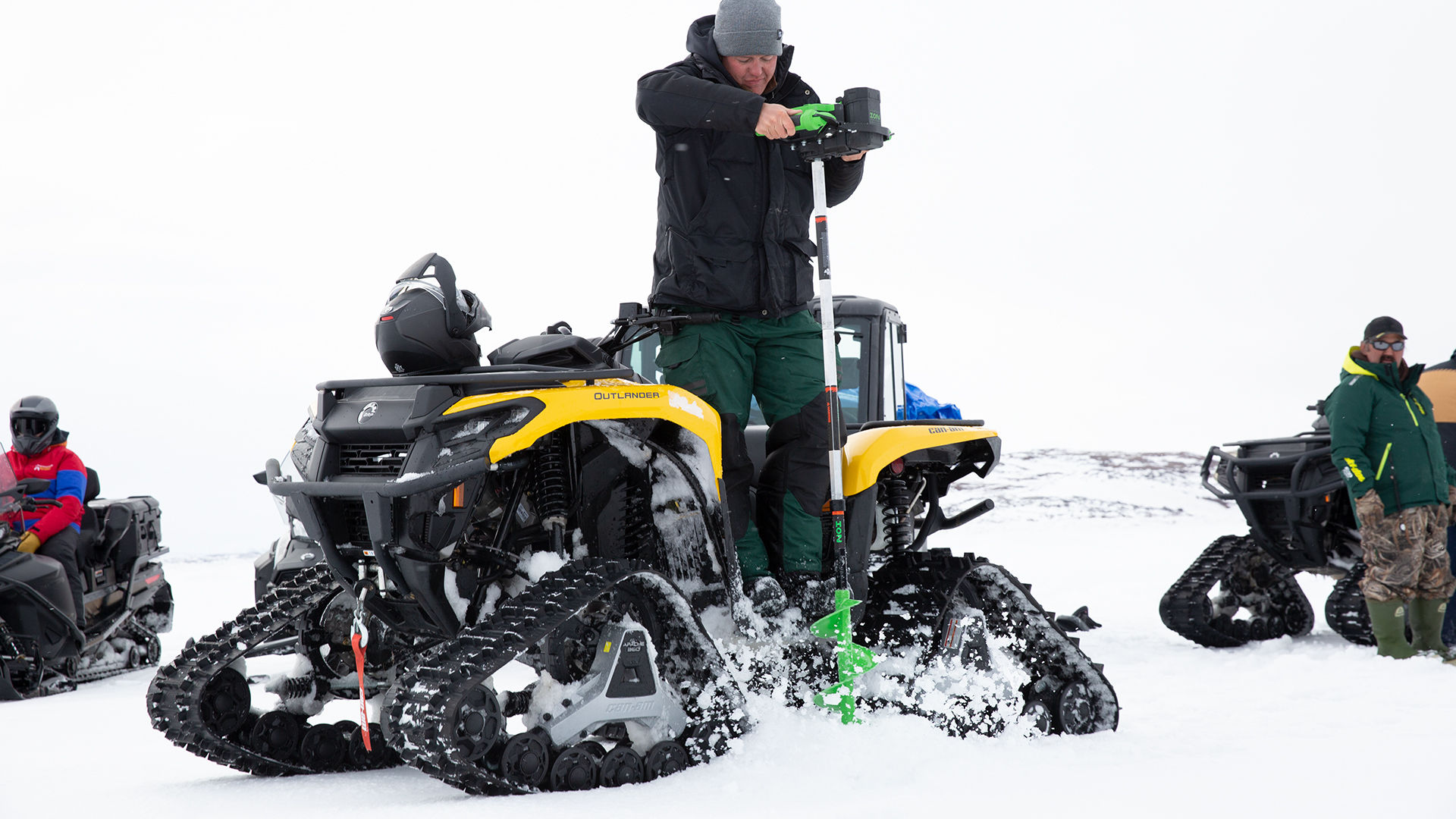 A man drilling a hole in the ice near his Can-Am ATV equipped with track system