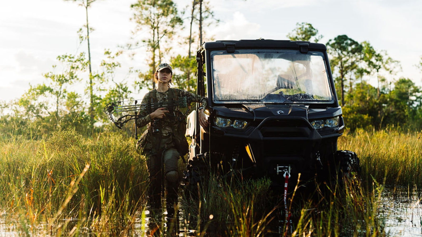 A woman with a crossbow placed next to her Can-Am side-by-side vehicle for hunting