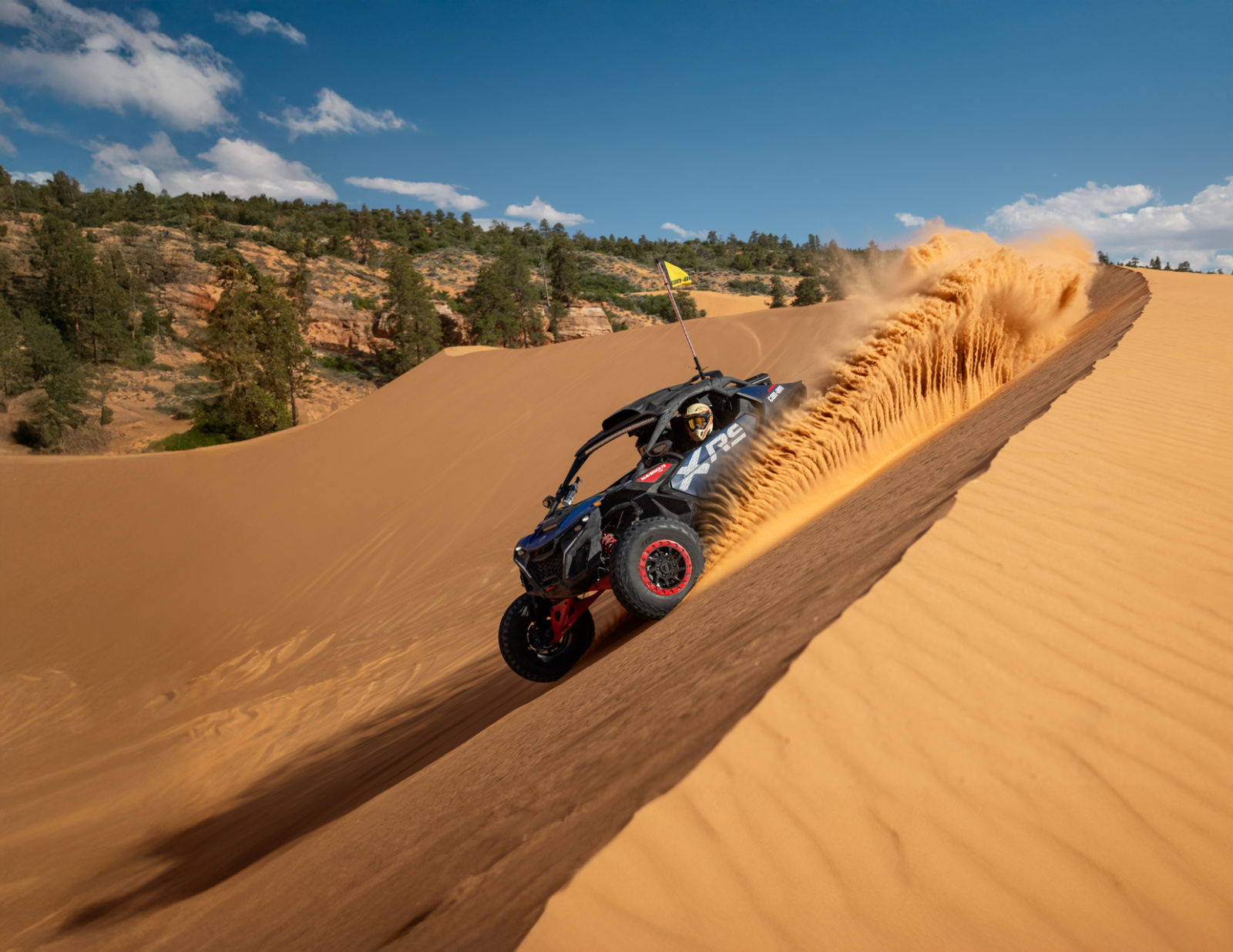 A Can-Am Maverick R X RS 2025 rolling down a sand dune on a sunny day