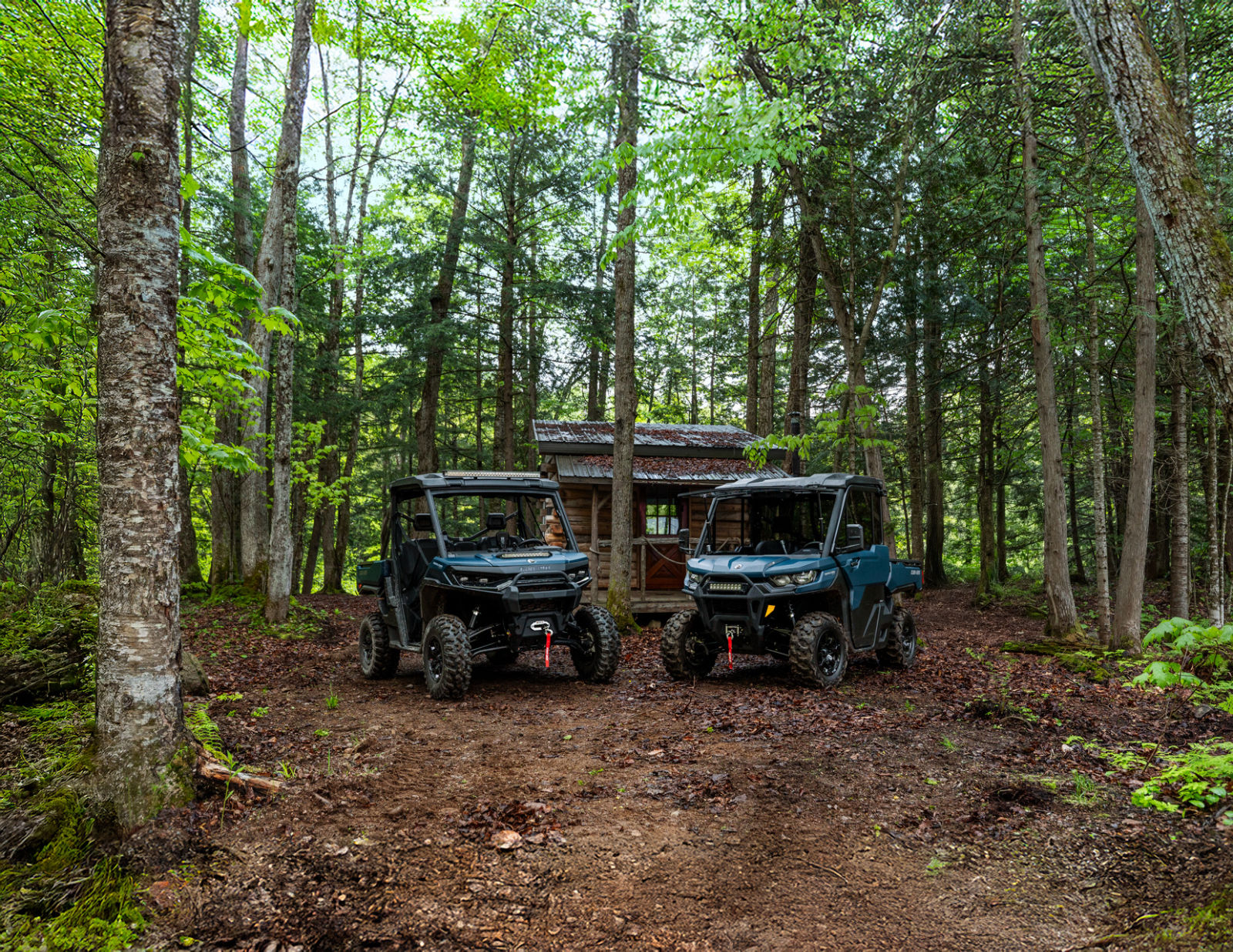 Two 2026 Can-Am Defender parked in a forest near a cabin