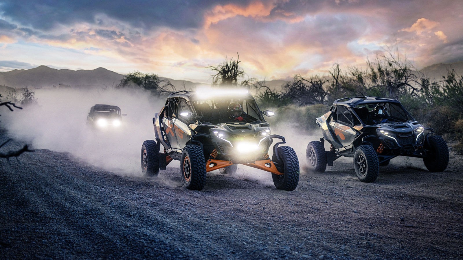 Three Can-Am Maverick side-by-side vehicles crossing the desert at dusk with their headlights on