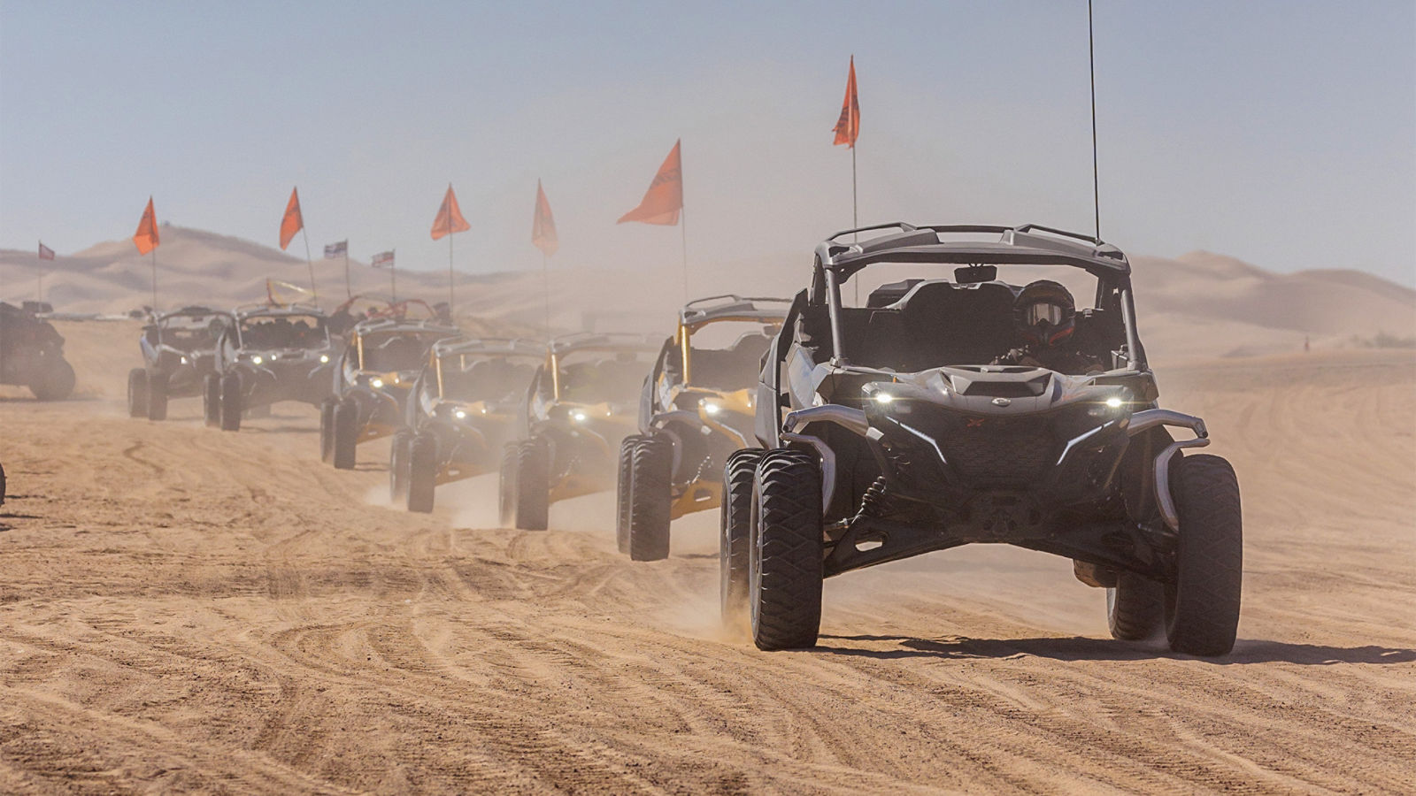 A convoy of Can-Am Maverick SxS with orange safety flags driving across desert sand dunes