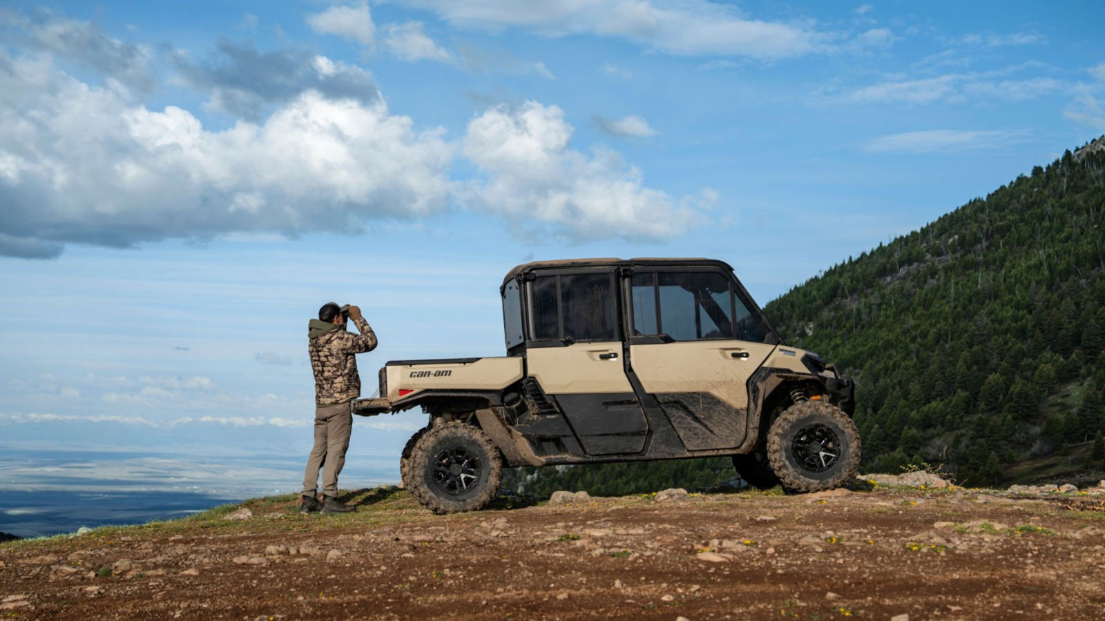 A hunter in camouflage standing by a 2027 Can-Am Defender XU side-by-side looking through binoculars