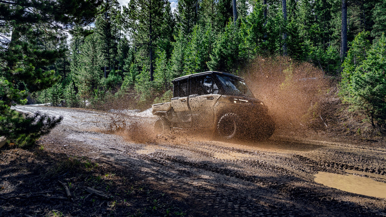 A 2027 Can-Am Defender side-by-side splashing through a deep mud puddle on a dirt forest trail