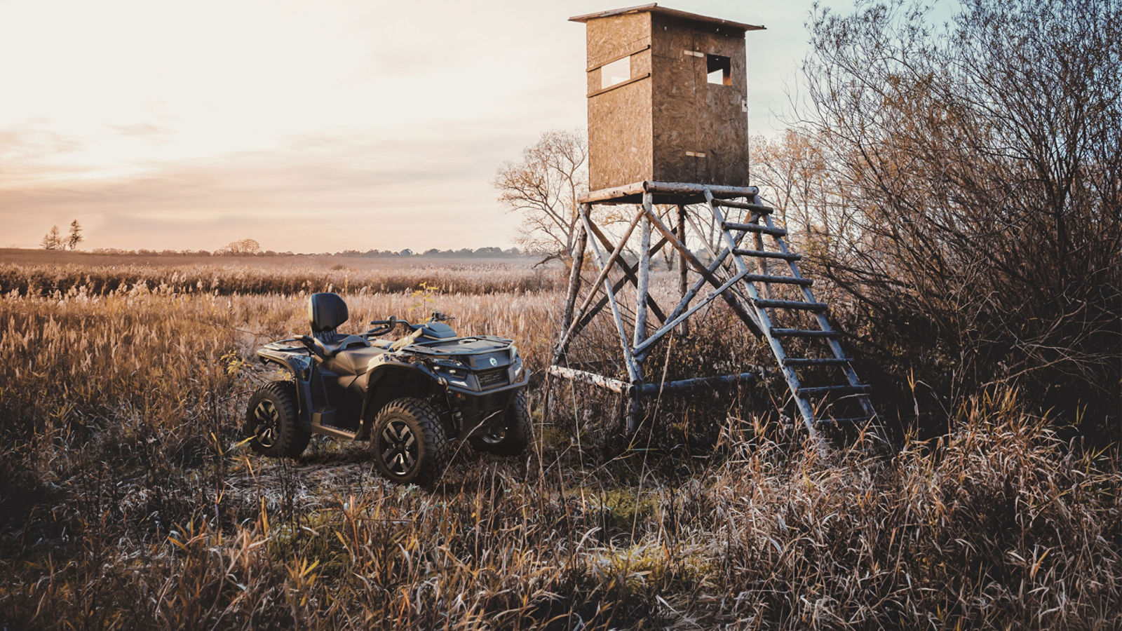 A 2027 Can-Am Outlander ATV parked next to an elevated wooden hunting blind in an open field