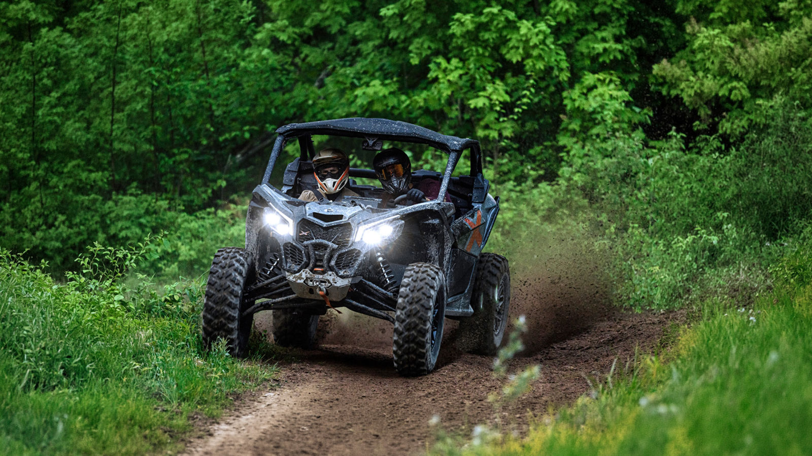 Two riders on a Can-Am Maverick Trail side-by-side on a lush green mountain trail