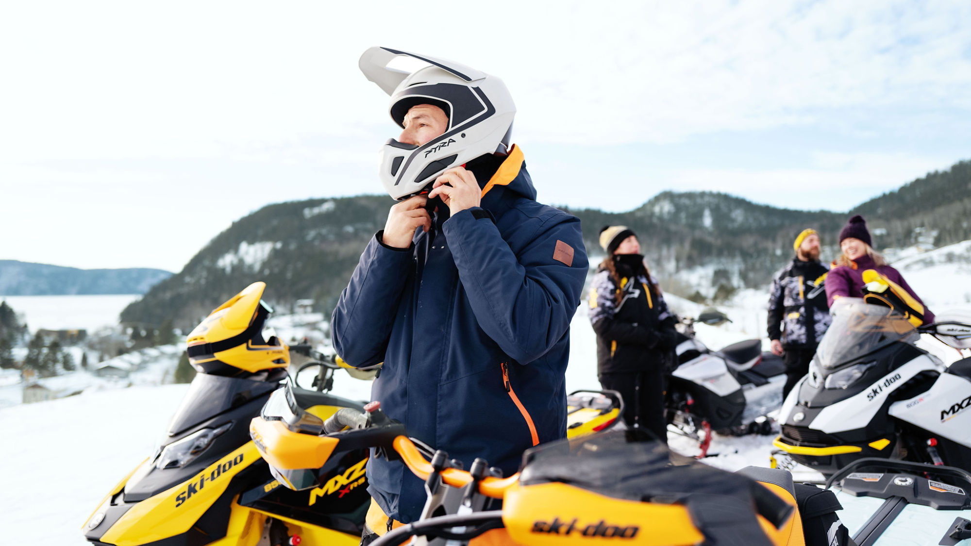 Man putting on his snowmobile helmet with his friends on the back