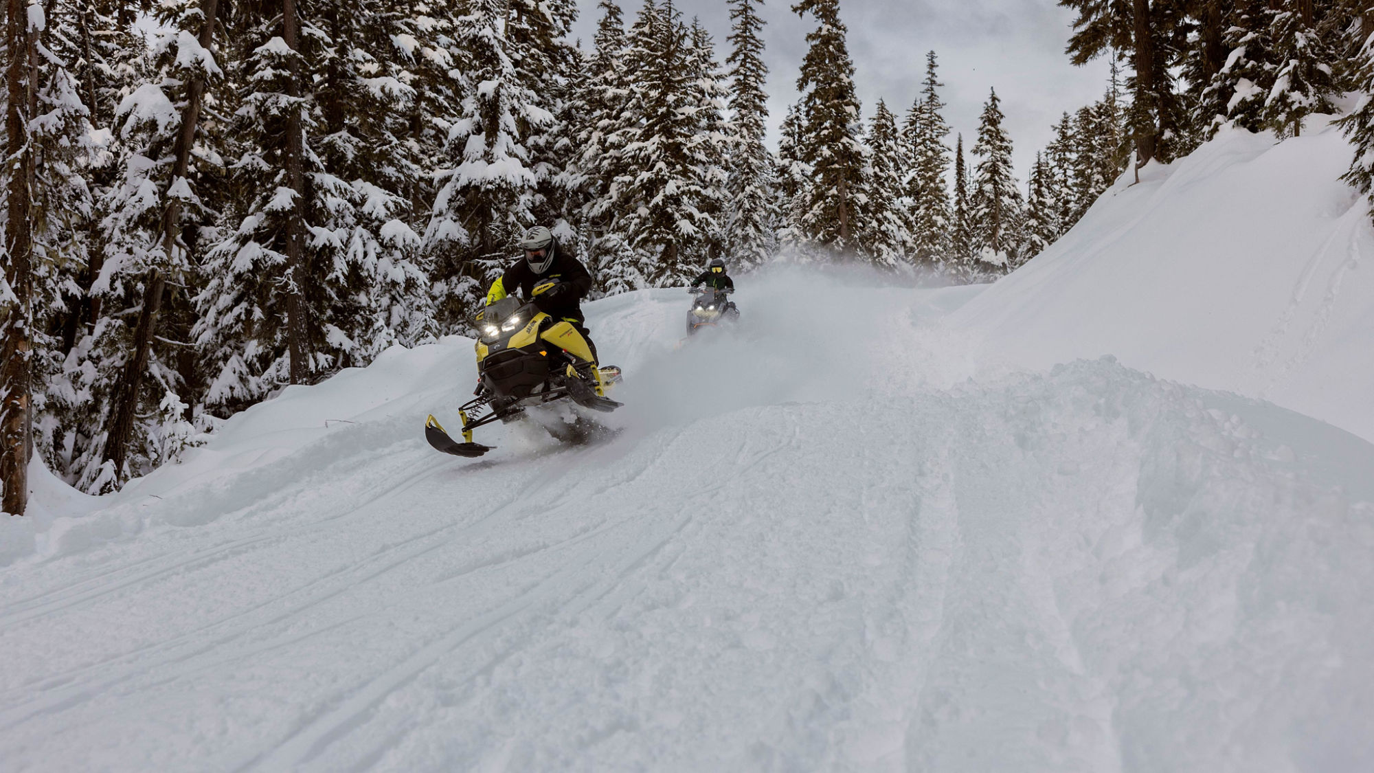 Två 2025 Ski-Doo Crossover snöskotrar kör i hög hastighet i en snöig skog