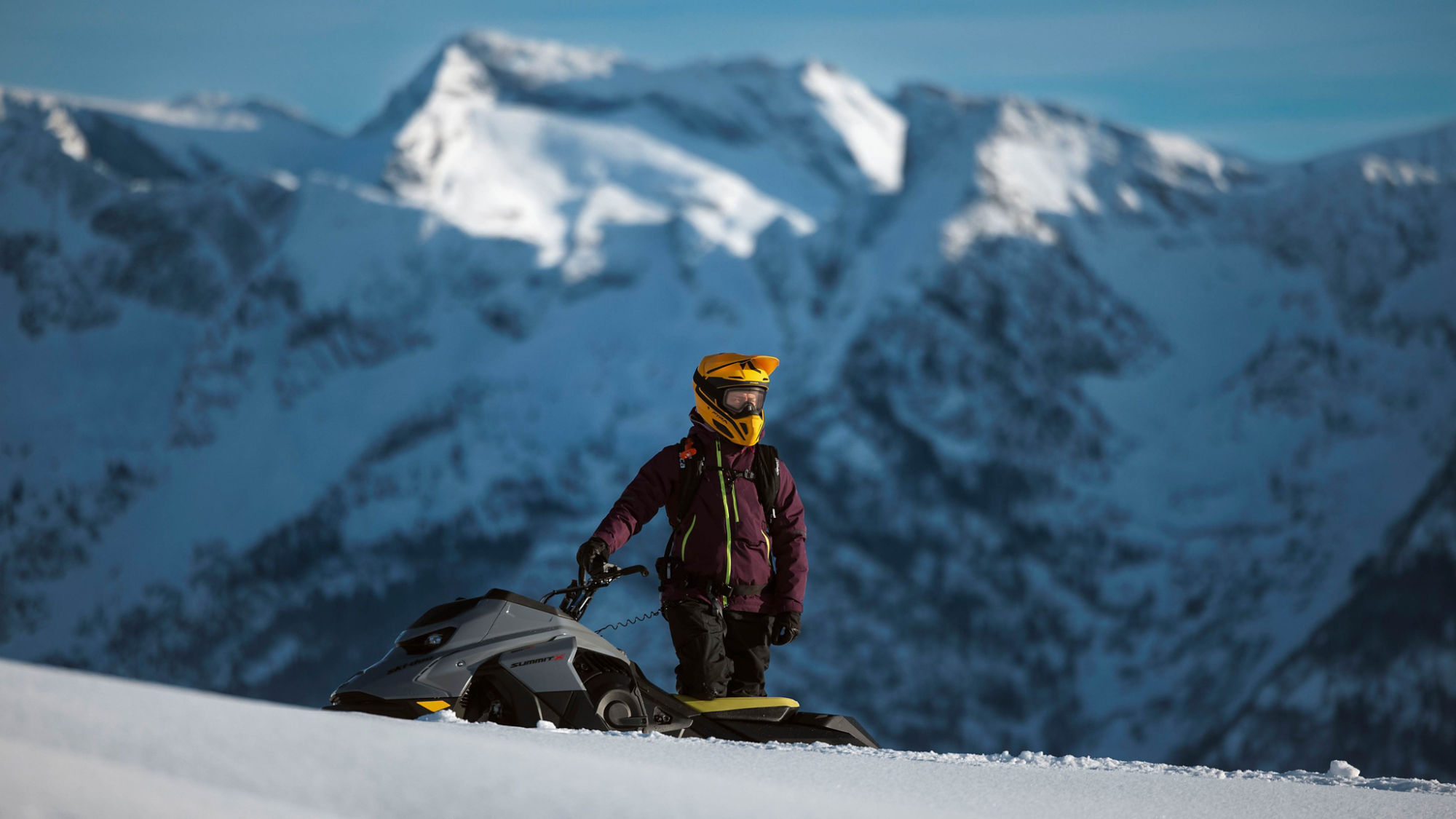 Snowmobiler standing next to his 2026 Ski-Doo Summit X snowmobile on a snowy mountain.