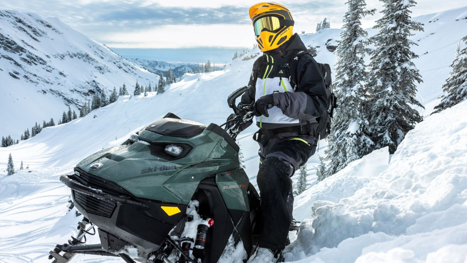 A rider beside a Ski‑Doo Summit X snowmobile on a snowy mountain slope with evergreen trees.