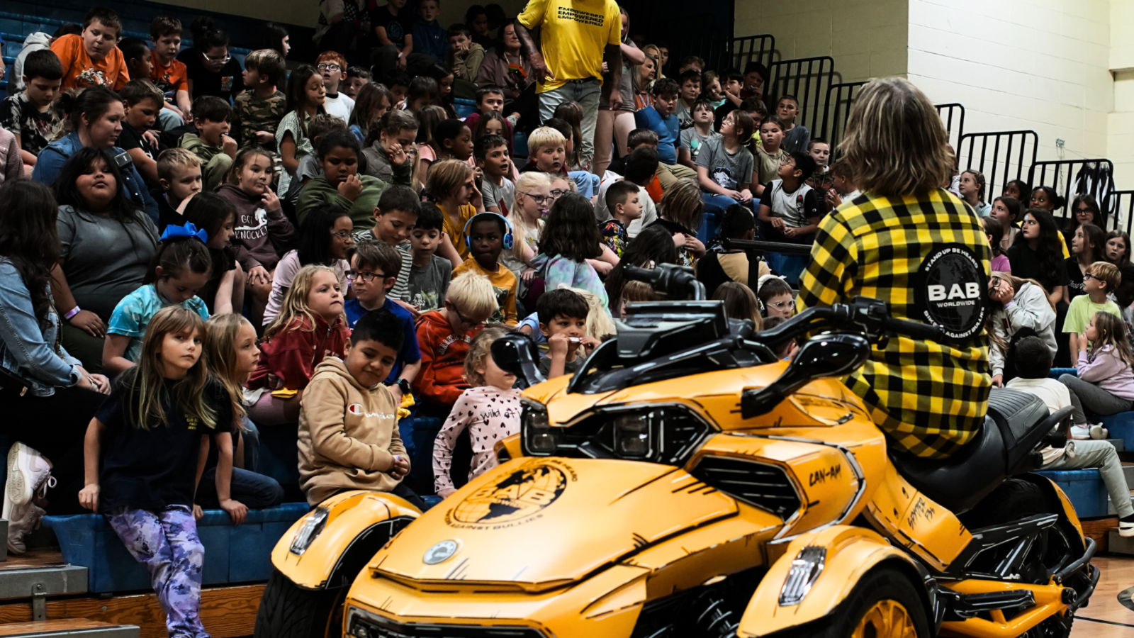 Hombre sentado en un vehículo de tres ruedas amarillo hablando con un grupo de niños.